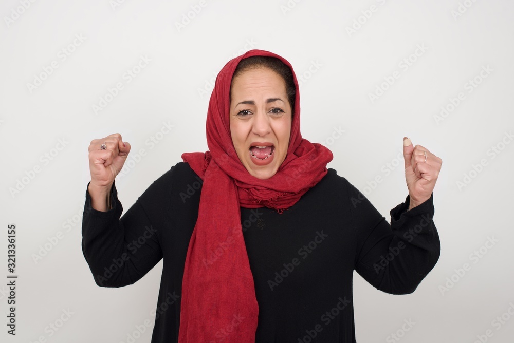 Waist up portrait of strong successful determined muslim female winner in casual t-shirt raising arms, clenching fists, exclaiming with joy and excitement. Victory, success and achievement concept. Waist up portrait of strong successful determined muslim female winner in casual t-shirt raising arms, clenching fists, exclaiming with joy and excitement. Victory, success and achievement concept.