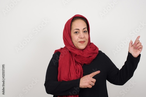 Pretty muslim woman with positive expression, dressed casually, indicates with fore finger at blank copy space for your promotional text or advertisement. Adorable woman poses indoor alone