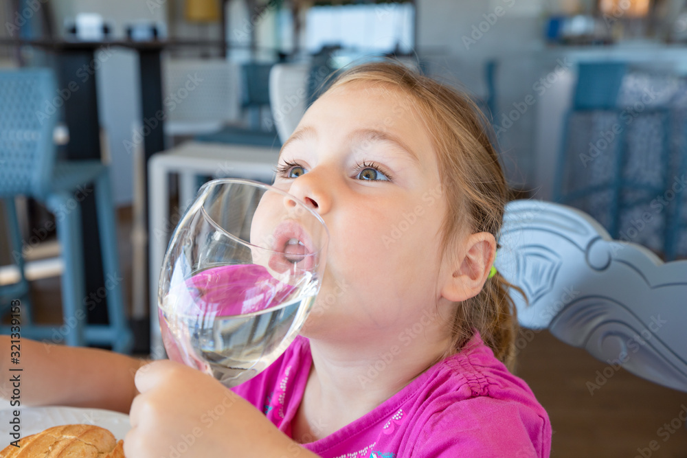 portrait of four years old blonde little girl with pink shirt drinking water from cup, sitting indoor in restaurant portrait of four years old blonde little girl with pink shirt drinking water from cup, sitting indoor in restaurant