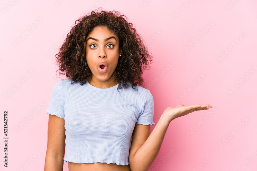 Young african american woman against a pink background impressed holding copy space on palm. Young african american woman against a pink background impressed holding copy space on palm.