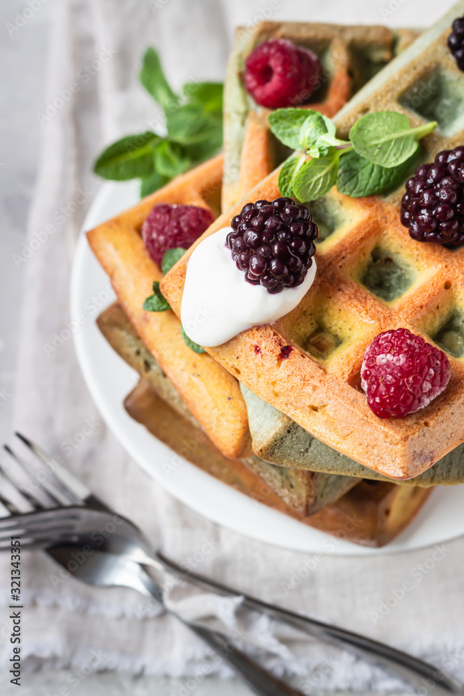 A stack of homemade Belgian waffles with frozen berries, mint and natural greek yogurt on a white plate, light grey stone background. Top view. A stack of homemade Belgian waffles with frozen berries, mint and natural greek yogurt on a white plate, light grey stone background. Top view.