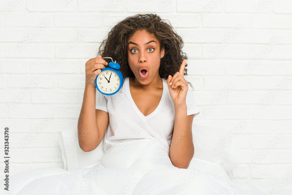 Young african american woman sitting on the bed holding an alarm clock having some great idea, concept of creativity. Young african american woman sitting on the bed holding an alarm clock having some great idea, concept of creativity.
