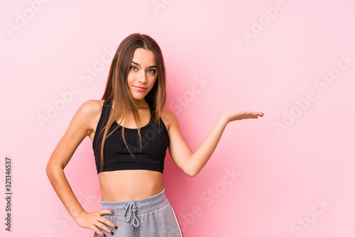 Young caucasian fitness woman posing in a pink background showing a copy space on a palm and holding another hand on waist.