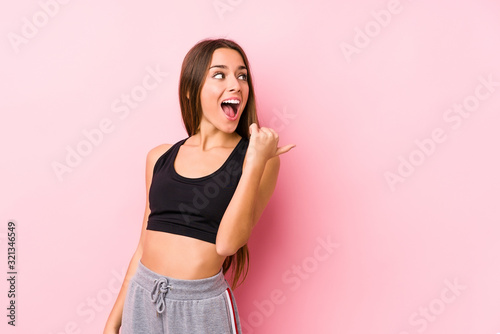 Young caucasian fitness woman posing in a pink background points with thumb finger away, laughing and carefree.