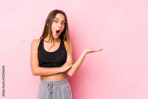 Young caucasian fitness woman posing in a pink background impressed holding copy space on palm.