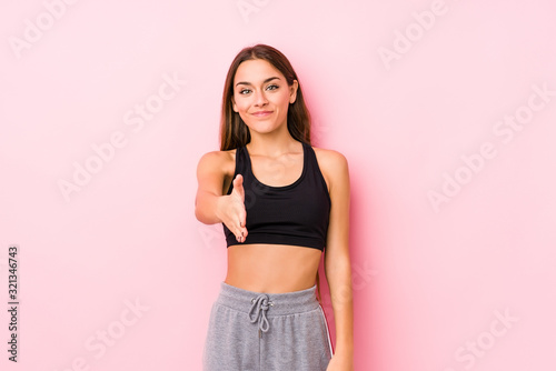 Young caucasian fitness woman posing in a pink background stretching hand at camera in greeting gesture.