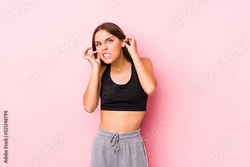 Young caucasian fitness woman posing in a pink background covering ears with hands. © Asier