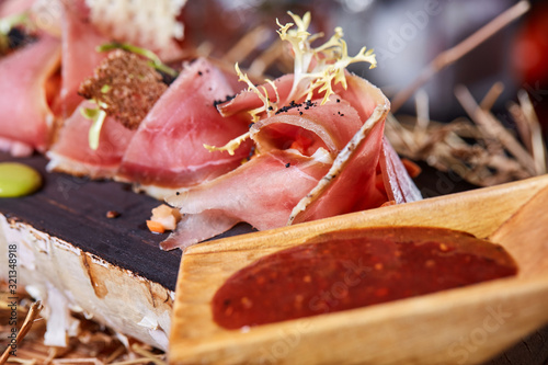Antipasto on wooden plate close up. Cold smoked meat plate with tomato hot sauce, sliced ham, prosciutto, bacon. Appetizer on wooden tray cut tree sawed imitation.