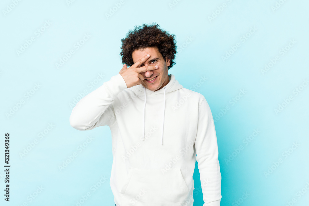 Young sporty man against a blue background blink at the camera through fingers, embarrassed covering face. Young sporty man against a blue background blink at the camera through fingers, embarrassed covering face.
