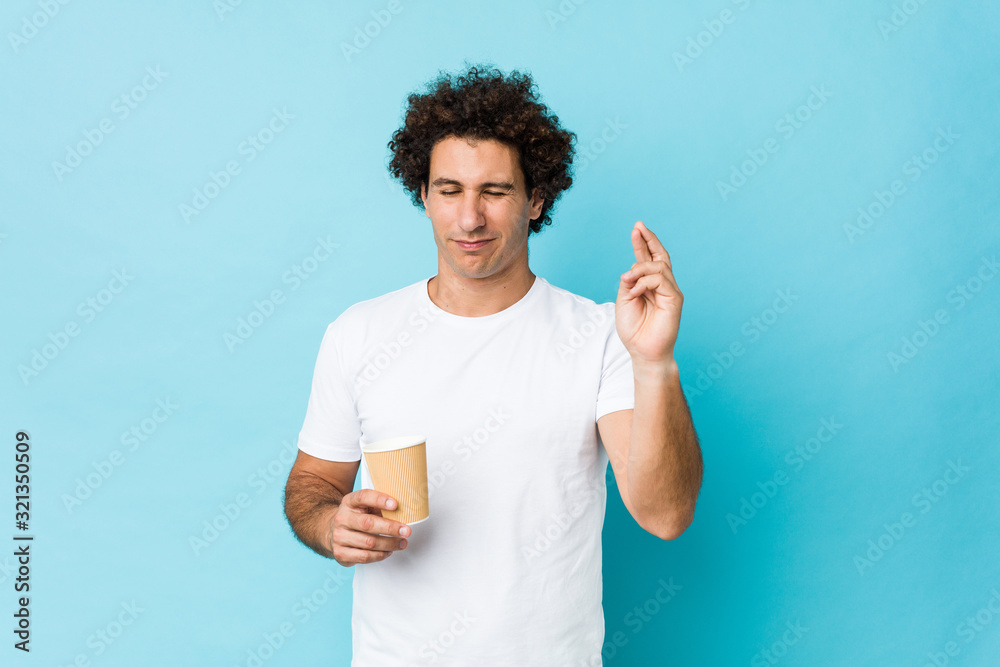 Young caucasian curly man holding a takeaway coffee crossing fingers for having luck Young caucasian curly man holding a takeaway coffee crossing fingers for having luck