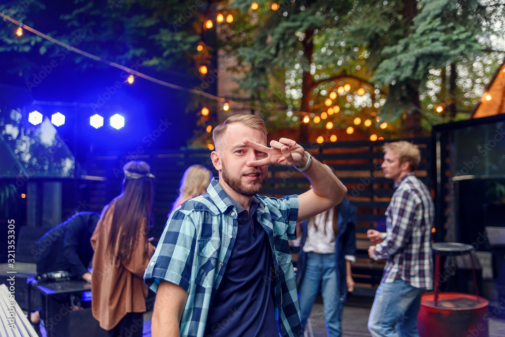 Handsome 30s caucasian young man dancing on near the camera on the background of his friends at the party in cozy evening garden.