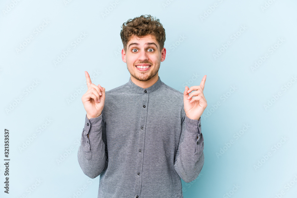 Young blond curly hair caucasian man isolated indicates with both fore fingers up showing a blank space. Young blond curly hair caucasian man isolated indicates with both fore fingers up showing a blank space.