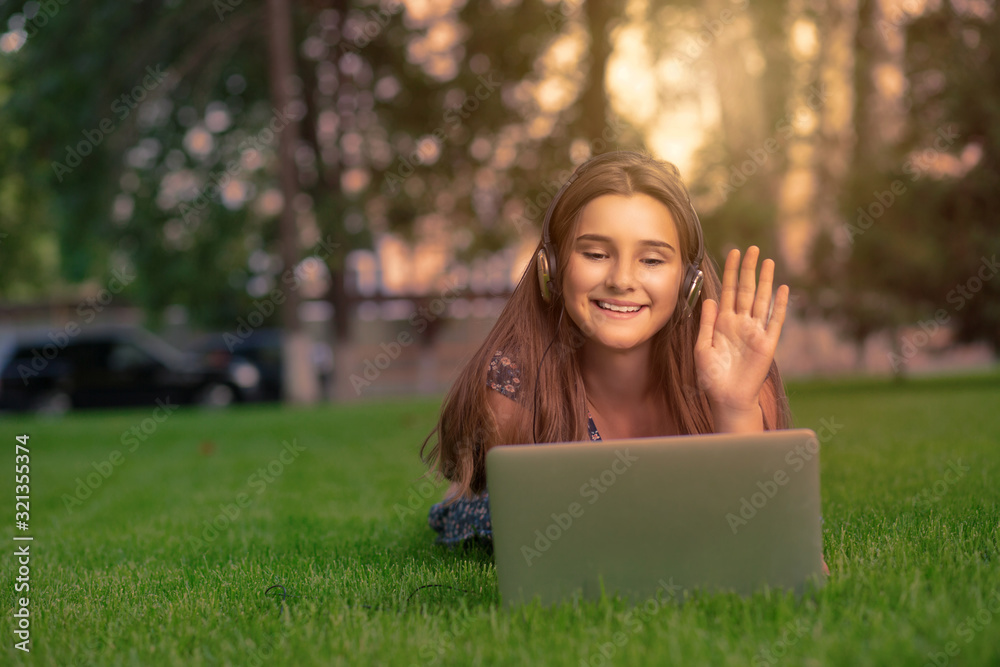 Hi there. Happy young cute student woman with smartphone headset on head and computer looking at screen greeting someone on video chat web conference lying on green grass lawn in park. Hi there. Happy young cute student woman with smartphone headset on head and computer looking at screen greeting someone on video chat web conference lying on green grass lawn in park.