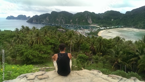 Man is sitting on top of a mountain and enjoys the view of the island's landscape with a lot of Palm trees, Ocean, beach and mountains