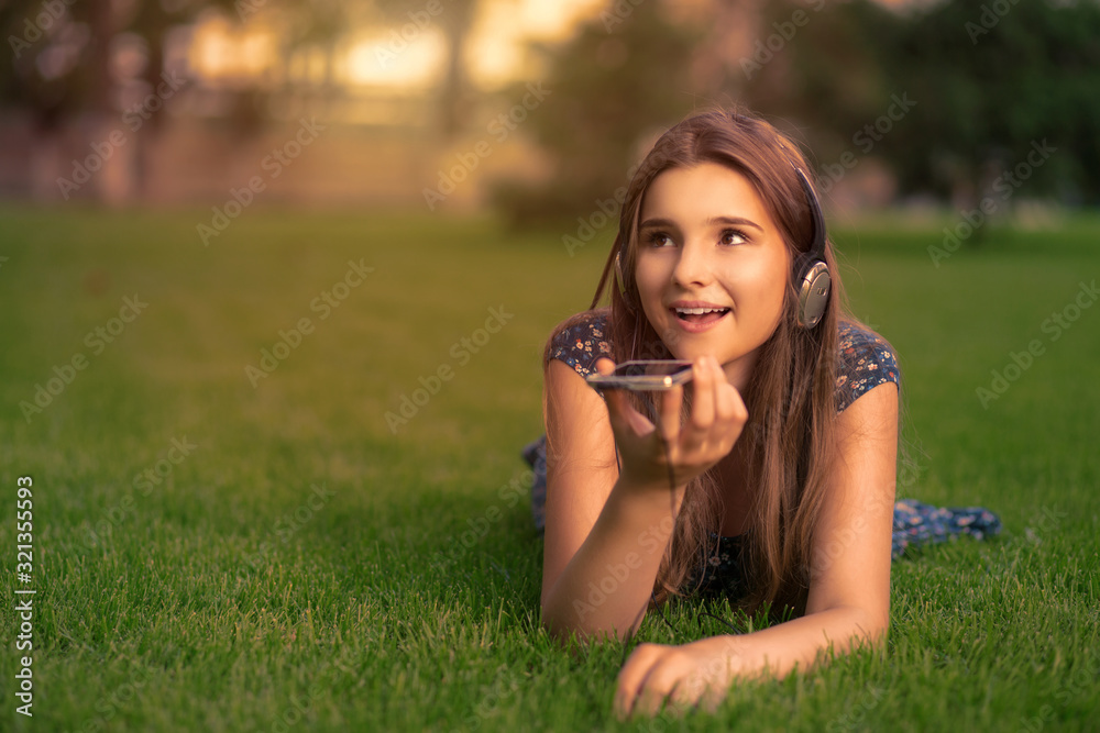 Student using voice recognition with a smart phone to record notes sitting on the grass in a park with headphones on head