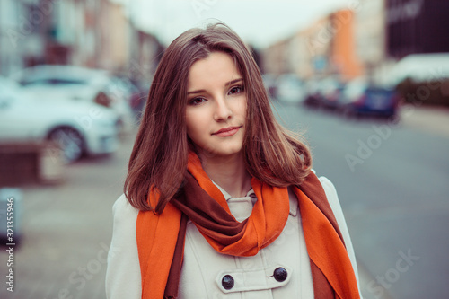 Attractive serious young Vietnamese woman in beige jacket coat and vivid red scarf isolated in an urban street looking at the camera you, close up view of her face and shoulders. Mixed race model