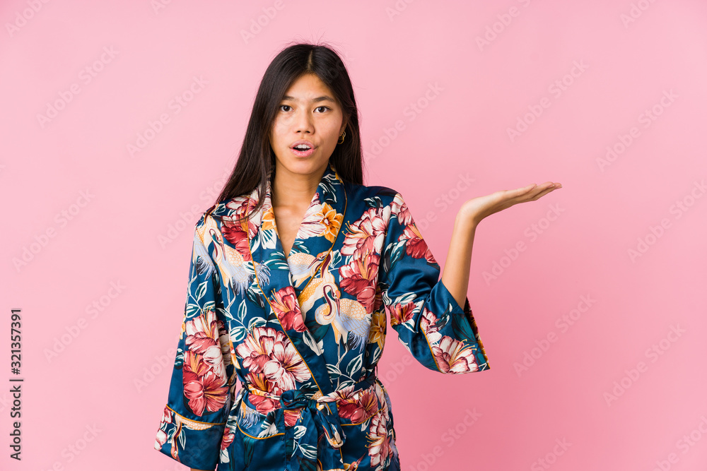 Young asian woman wearing a kimono pajamas impressed holding copy space on palm. Young asian woman wearing a kimono pajamas impressed holding copy space on palm.