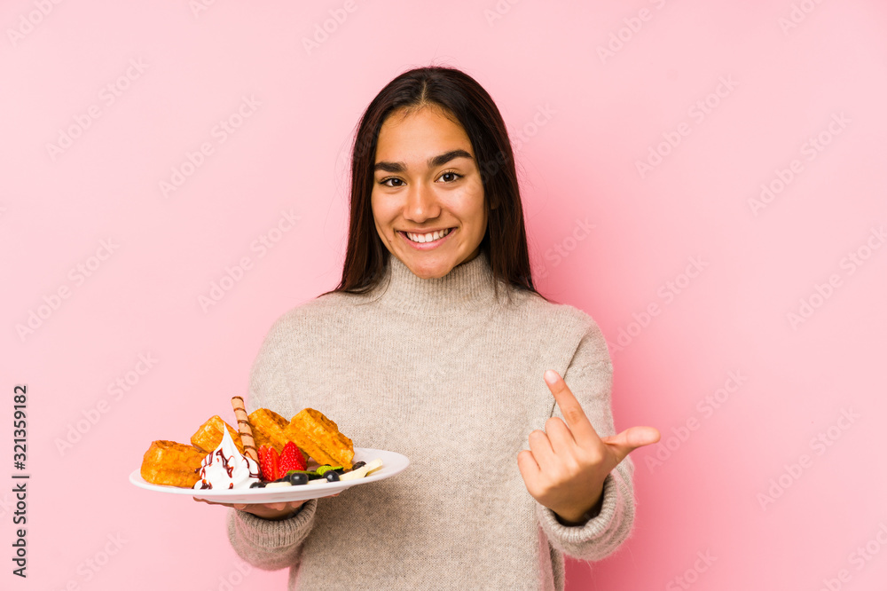 Young asian woman holding a waffle isolated pointing with finger at you as if inviting come closer.