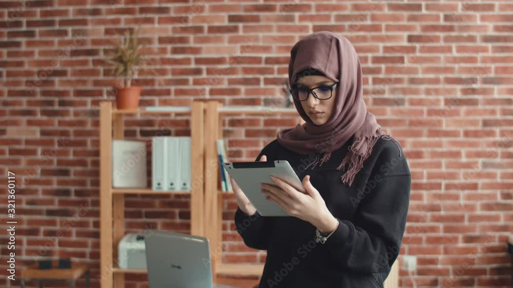 A Muslim woman is typing on a digital tablet and looking at the camera. A young woman in a hijab works on an electronic tablet and looks at the camera smiling