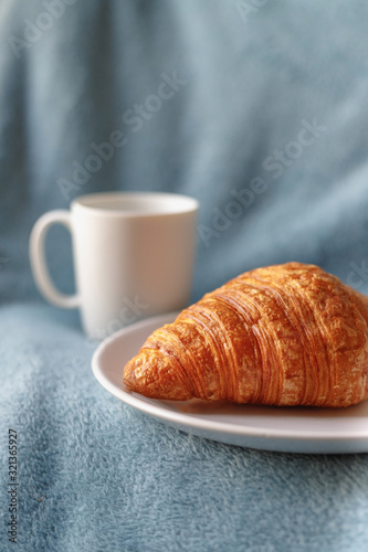 Tasty croissant on plate near coffee in white cup in bed. Vertical close up shot. Cozy morning at home.