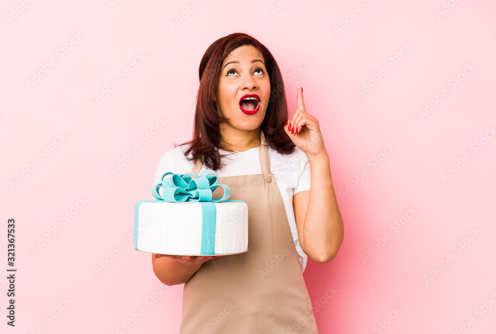 Middle age latin woman holding a cake isolated on a pink background pointing upside with opened mouth. Middle age latin woman holding a cake isolated on a pink background pointing upside with opened mouth.