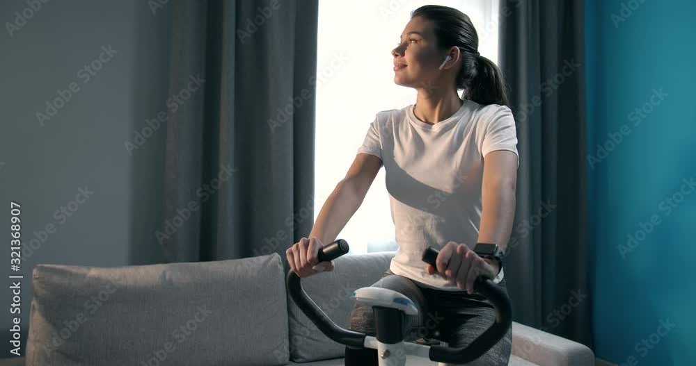 Motivated young girl in sportswear doing exercise on bicycle at home and listening music on earphones. Charming brunette training everyday to stay fit and healthy Motivated young girl in sportswear doing exercise on bicycle at home and listening music on earphones. Charming brunette training everyday to stay fit and healthy