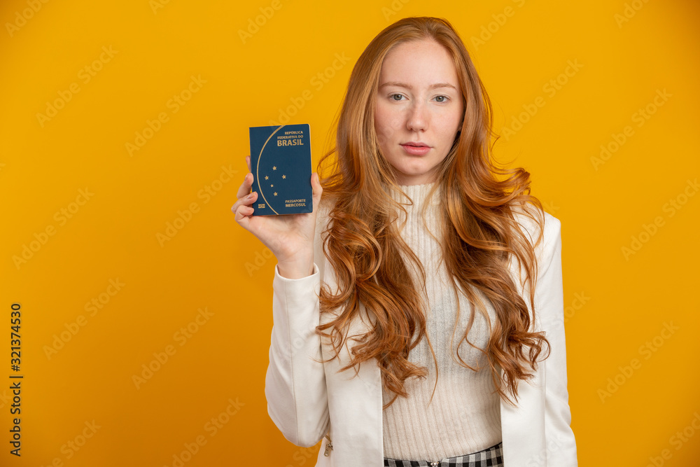 Beautiful redhead curly-haired girl in travel. Next trip. Girl holding Brazilian passport. On yellow. Beautiful redhead curly-haired girl in travel. Next trip. Girl holding Brazilian passport. On yellow.
