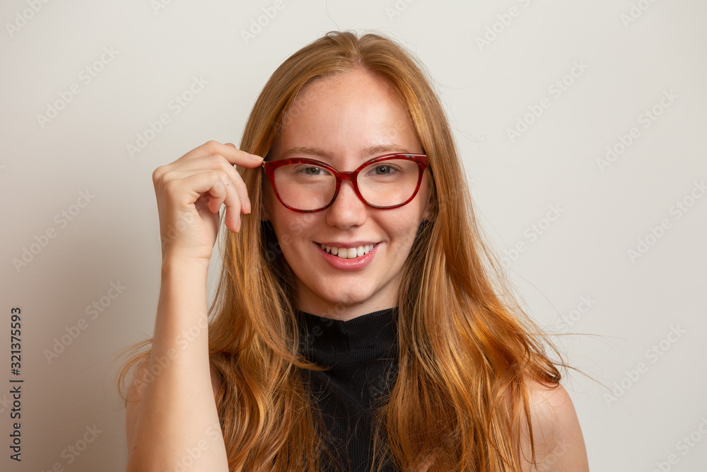 Beautiful young redhead girl in eyeglasses is looking at camera and smiling, on gray background. Beautiful young redhead girl in eyeglasses is looking at camera and smiling, on gray background.
