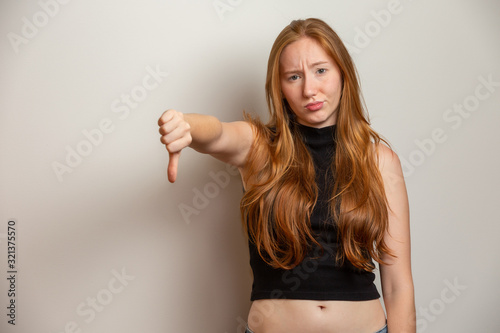 Unhappy redhead woman giving thumbs down gesture looking with negative expression and disapproval. Yellow background.