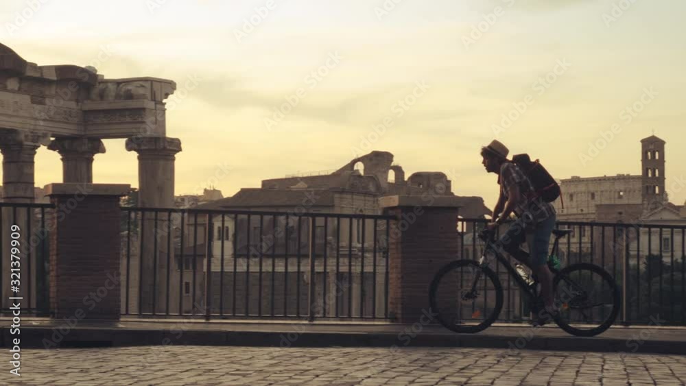 Young man tourist with backpack and bike cycling at Roman Forum at sunrise. Historical imperial Foro Romano in Rome, Italy from panoramic point of view.