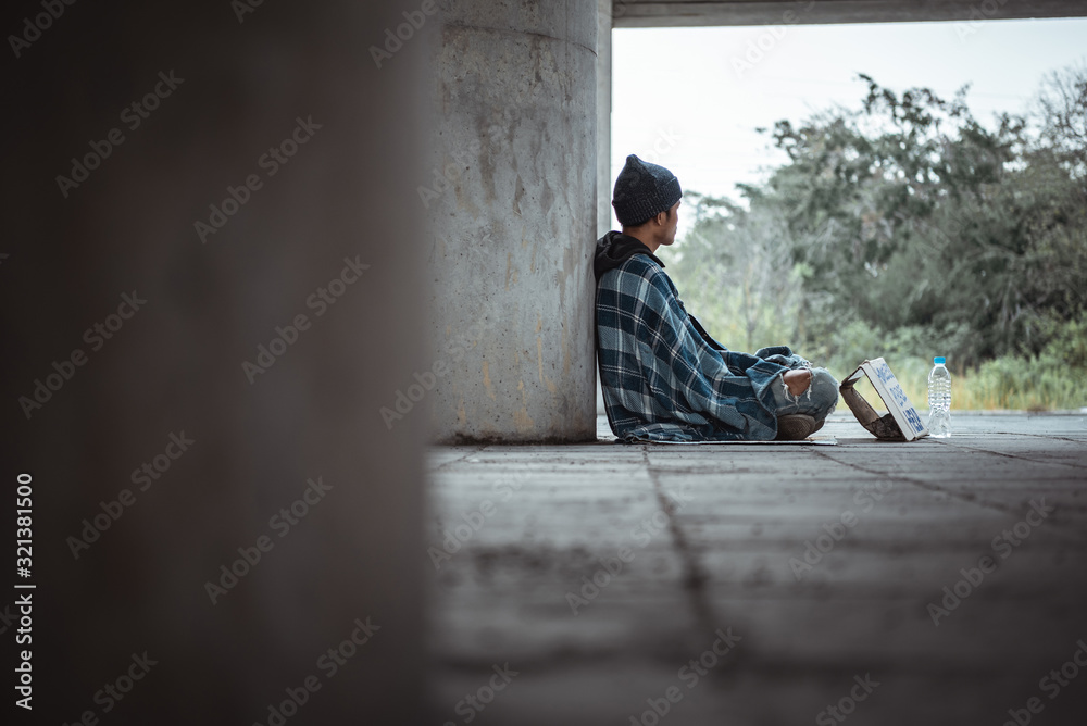 Fototapeta premium Homeless man sitting on the street in the shadow of the building and begging for help and money.