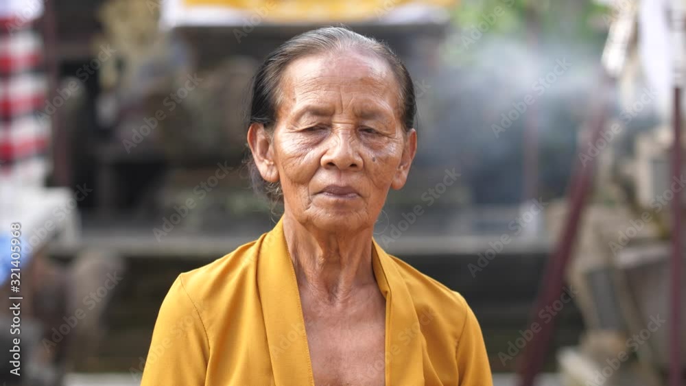 Elderly Asian woman stands in courtyard of Hindu temple in Bali with smoke from incense blowing in background. Old, weathered Balinese lady in bright yellow smiles at camera. Elderly Asian woman stands in courtyard of Hindu temple in Bali with smoke from incense blowing in background. Old, weathered Balinese lady in bright yellow smiles at camera.