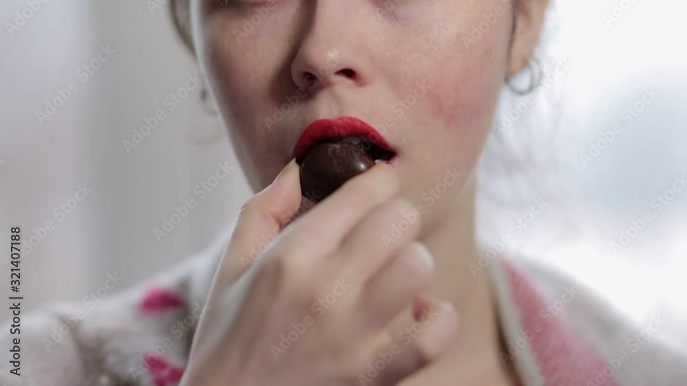 A young woman with red lips brings a chocolate candy to her mouth, bites and chews it. Lips close up