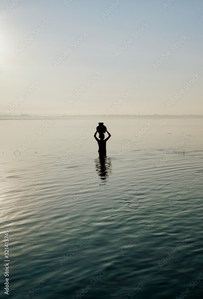 silhouette of a boy holding a bamboo jar above the head, standing in the water at sunrise