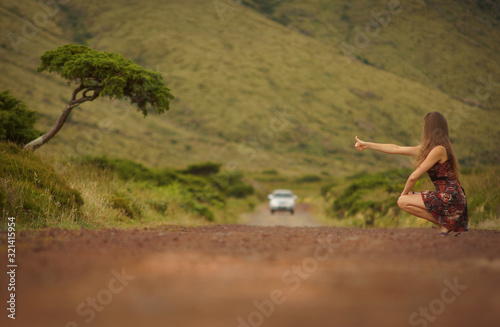 Young woman in dress hitching a ride on country road. Car on road, single tree on left side, green hills on background