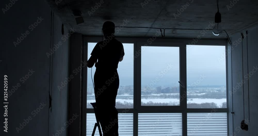 Silhouette of male electrician hold out electric wires in new apartment, stands on stepladder, fastens wires to ceiling, works in front of window with beautiful view of river, works with hands, winter Silhouette of male electrician hold out electric wires in new apartment, stands on stepladder, fastens wires to ceiling, works in front of window with beautiful view of river, works with hands, winter