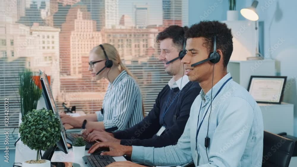 Portrait of smiling mixed-race man taking call in busy call center. In the background there are his colleagues also talking to clients on phone.