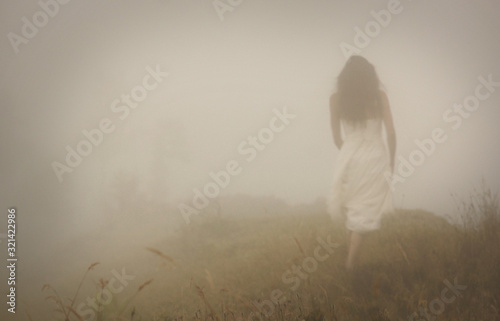 Woman in long white dress going in foggy mist. Blur discernible outline of figure. View from behind. Mistery landscape