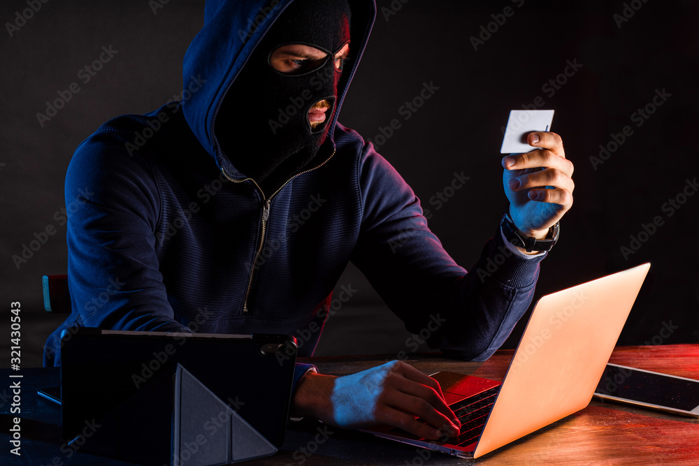 young caucasian hacker man in the hood of pullover sit holding bank card, going to crack algorithm to gain access to a computer system, want to withdraw money. isolated in dark room, black background young caucasian hacker man in the hood of pullover sit holding bank card, going to crack algorithm to gain access to a computer system, want to withdraw money. isolated in dark room, black background