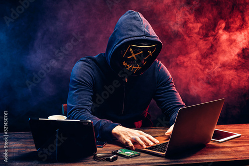 hacker man typing on laptop, hacking computer system. male in mask and pullover. unrecognizable incognito male sit in hood and try to breach the security of laptop system. neon smoke in background photo