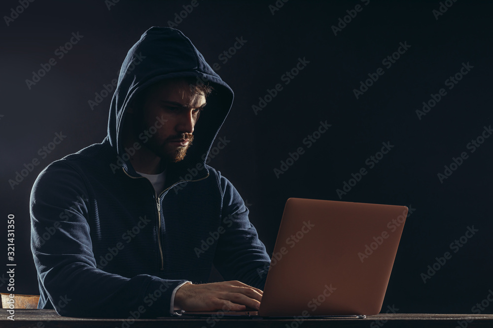 serious caucasian hacker male sit at table, look at screen of laptop studying computer system and typing password hacking bank account or online bullying, sit isolated in dark space, black background serious caucasian hacker male sit at table, look at screen of laptop studying computer system and typing password hacking bank account or online bullying, sit isolated in dark space, black background