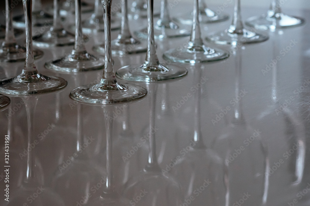 Many transparent high stalk glasses on the table, with beautiful reflections. A raw of empty wine glasses. Dim natural light from the window. Catering, holiday, cafe, restaurant glassware