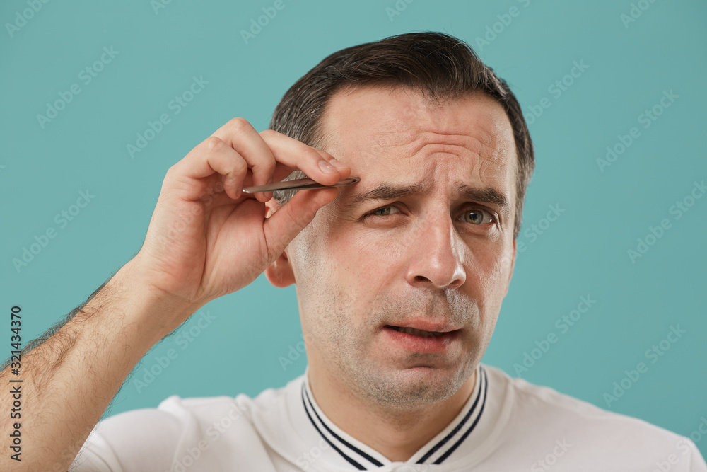 Head and shoulders portrait of wincing middle aged man plucking eyebrows standing against blue background Head and shoulders portrait of wincing middle aged man plucking eyebrows standing against blue background