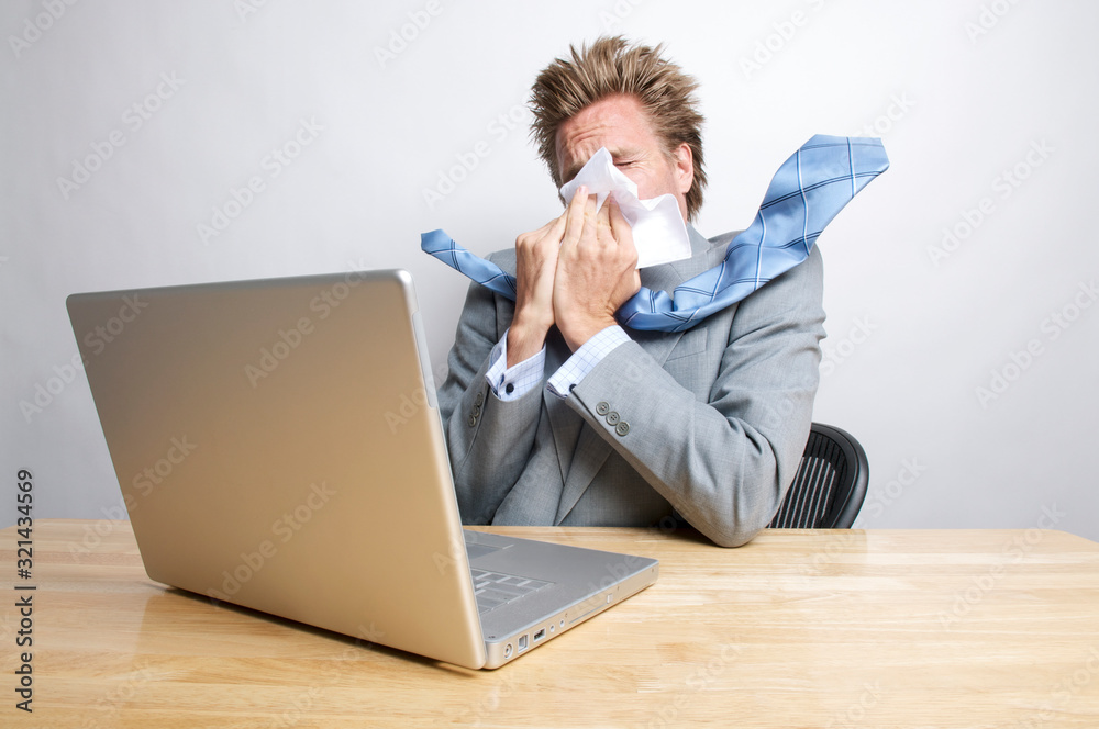 Sick businessman sneezing in front of his laptop computer sitting at his office desk Sick businessman sneezing in front of his laptop computer sitting at his office desk