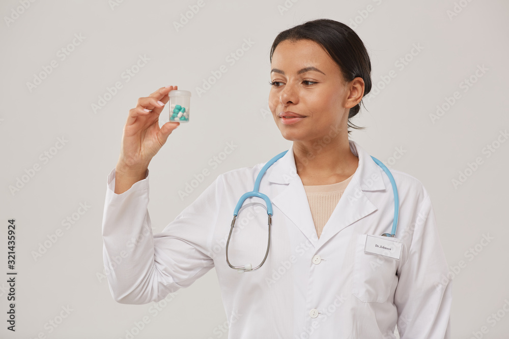 Waist up portrait of beautiful female doctor holding pill bottle while standing against white background, copy space Waist up portrait of beautiful female doctor holding pill bottle while standing against white background, copy space