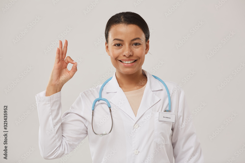 Waist up portrait of beautiful female doctor showing OK sign and smiling cheerfully at camera while standing against white background, copy space Waist up portrait of beautiful female doctor showing OK sign and smiling cheerfully at camera while standing against white background, copy space