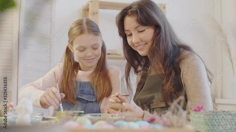 Medium shot of happy young woman and preteen girl suing brushes and painting on wooden Easter ornament Medium shot of happy young woman and preteen girl suing brushes and painting on wooden Easter ornament