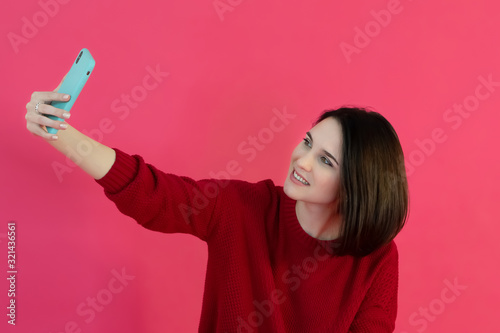 Beautiful young girl with short hair in a red sweater makes selfie on the phone on a bright pink background. Studio photo. © Luiza
