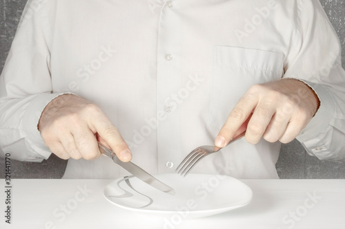 A man without a face sits at a table with a knife and a fork in his hands and eats imaginary food from an empty plate.