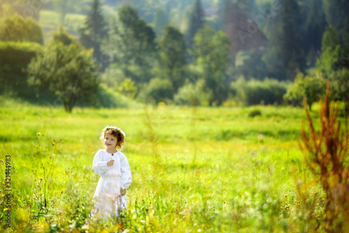 5 years old girl in white traditional embroidery ukrainian dress and chamomiles wreath on meadow in summer with Carpathian mountains on background. Travel tourism destination in Ukraine. Allergy time.